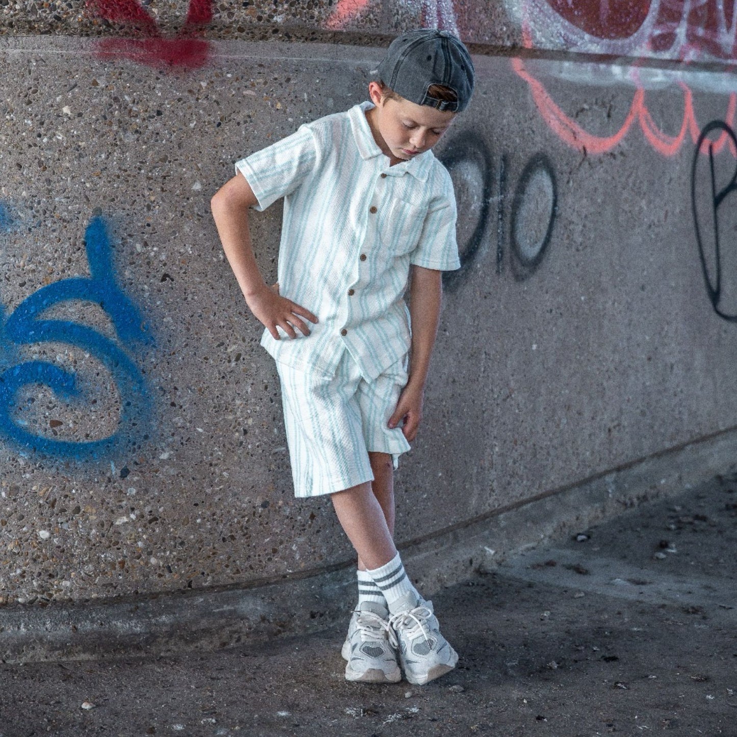 Kind met een grijs gewassen denim pet op achterstevoren, van voren gefotografeerd in een urban omgeving met graffiti.